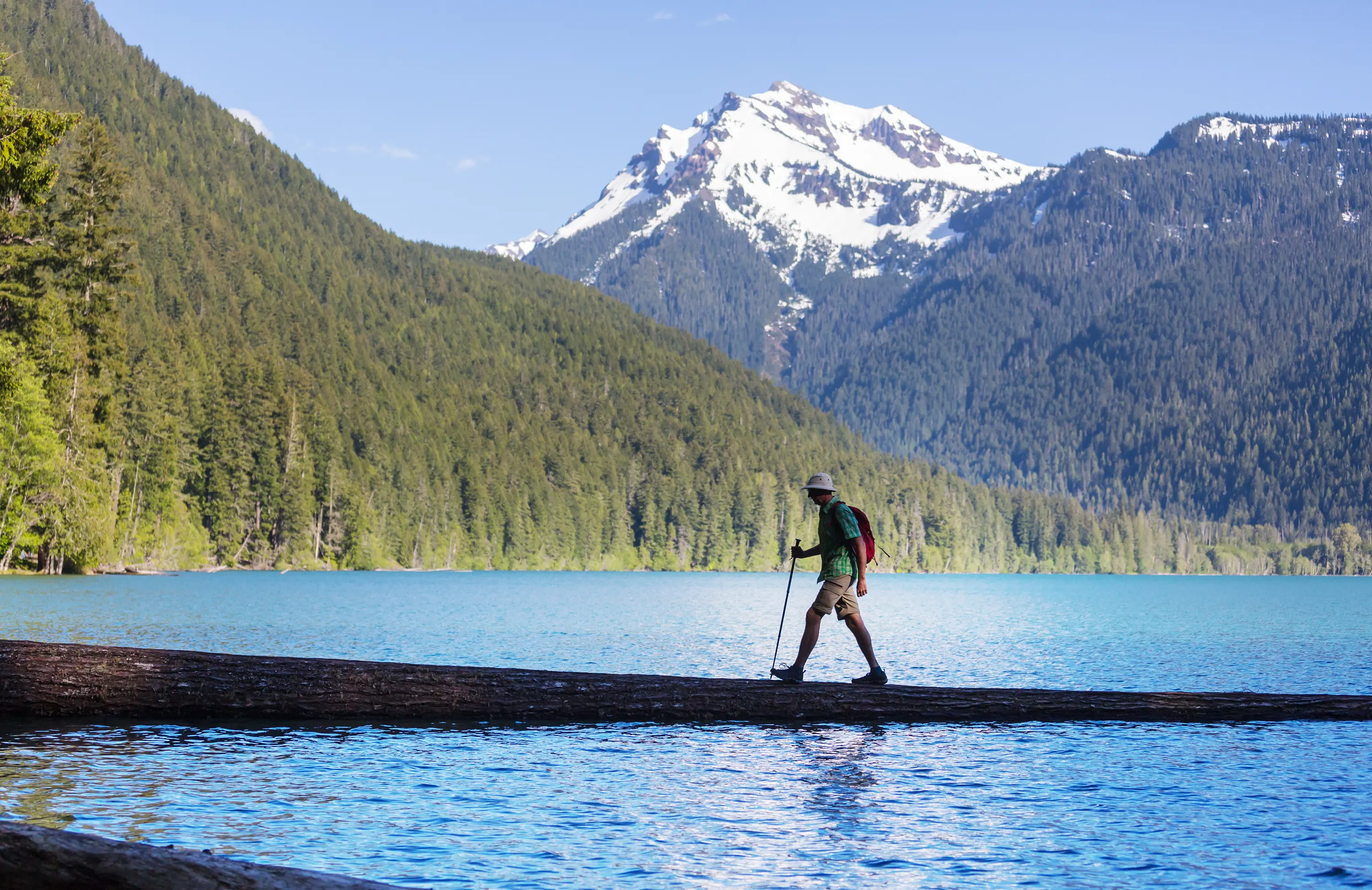 Caminhante cruza um tronco sobre um lago azul cercado por montanhas