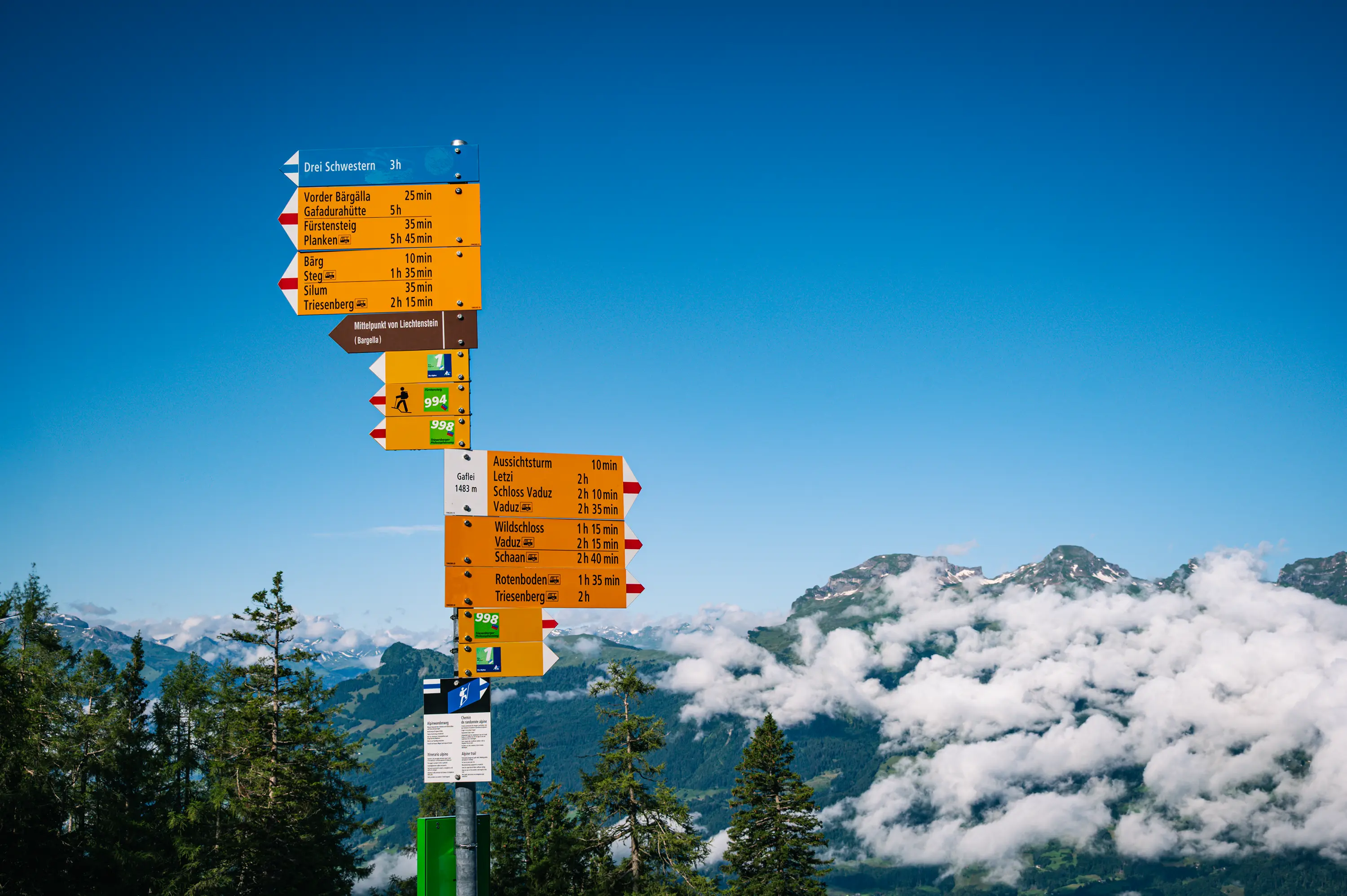 Poste de sinalização em Gaflei, Liechtenstein, com placas amarelas, placa azul para Drei Schwestern, números de rotas e indicação de altitude.